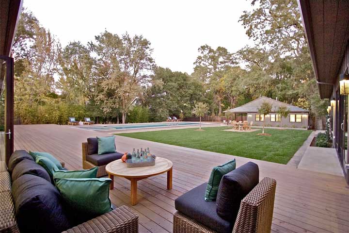 The spacious backyard features wooden flooring and comfortable sofas and chairs arranged around a table. Across the image, a sparkling pool adds to the outdoor oasis.