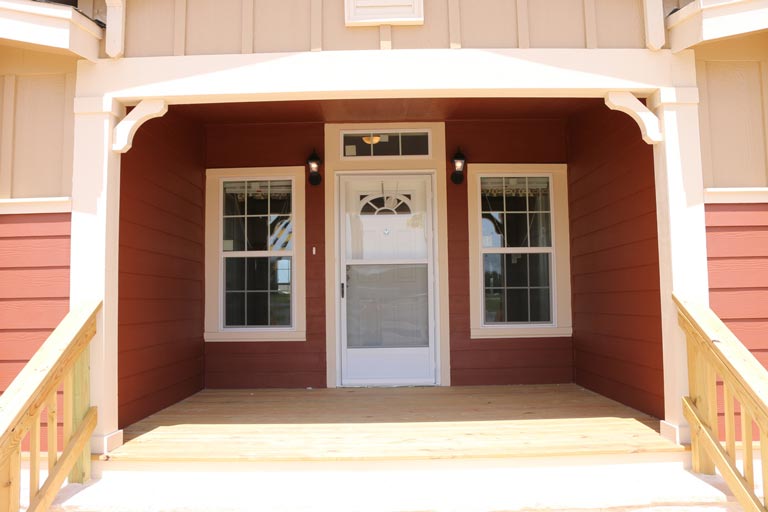 The image depicts a white wooden door on the front porch. The door is surrounded by two wooden windows on either side, and there are two light sconces mounted on the porch wall.