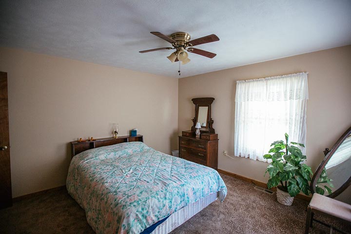 This bedroom is well-lit and inviting, and features a comfortable bed, a large window that allows natural light to pour in, and a ceiling fan to keep the air circulating.