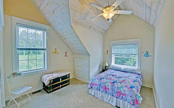 Another bedroom with a comfortable bed, dressed in white linens and a fluffy comforter. The room is painted in a soft hue, with large windows that lets in plenty of natural light.