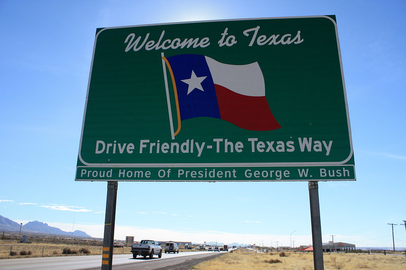 Welcome to Texas state sign in green next to a highway with a few cars in a background