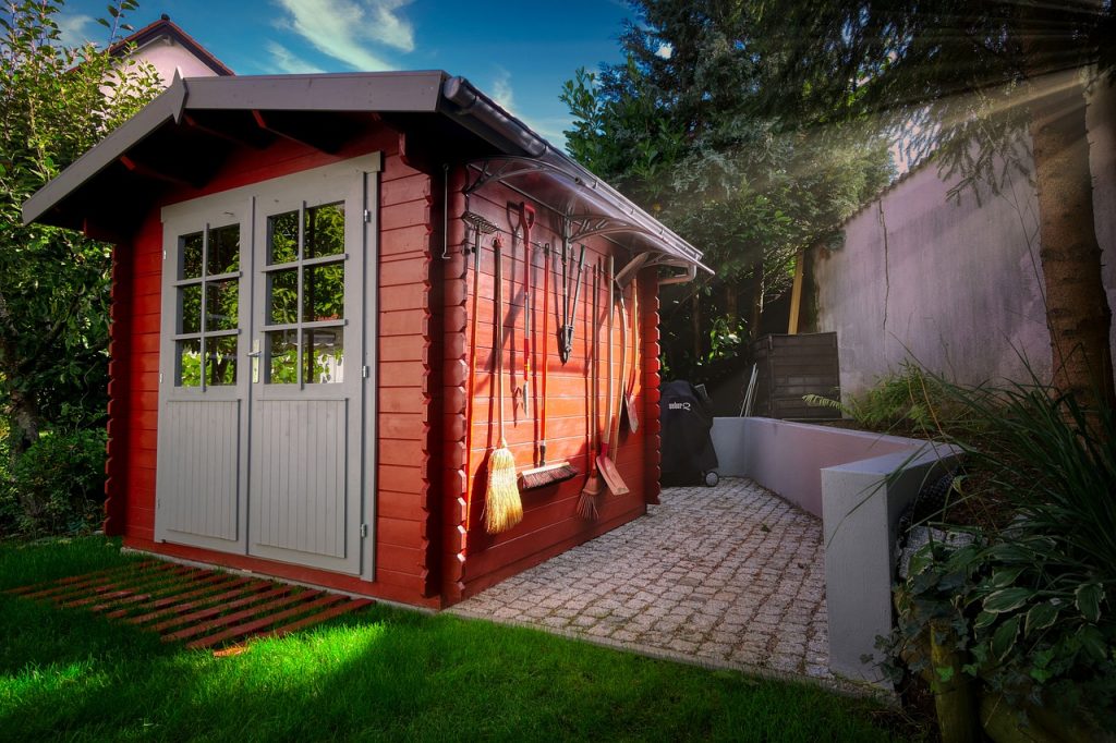 Red garden shed with white doors and gardening tools hooked on the side.
