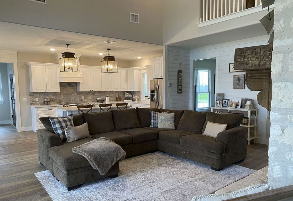 Living room and kitchen layout with a brown sectional sofa, light-gray rug,  ceiling lights, and white kitchen cabinets. 