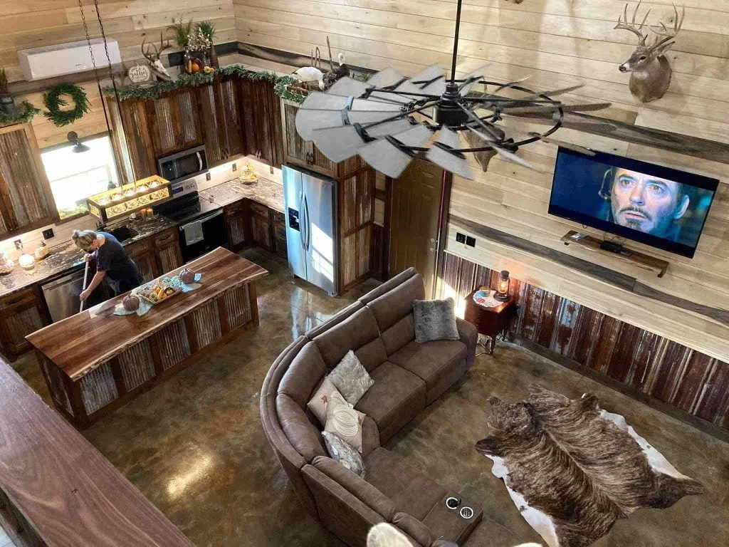 an interior of a sitting room in 40 x 60 barndominium from above the loft featuring corner sofa, metal kitchen finishes, tv, and a cow hide carpet