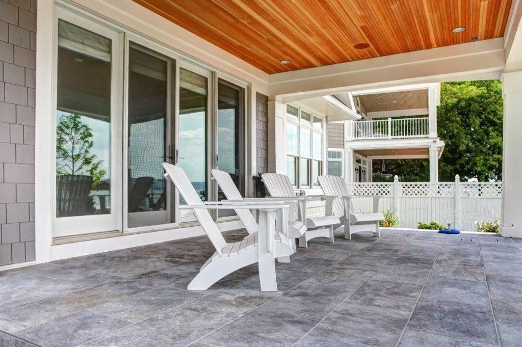 The covered porch with white lounge chairs and behind, glass doors that lead to the great room inside. 