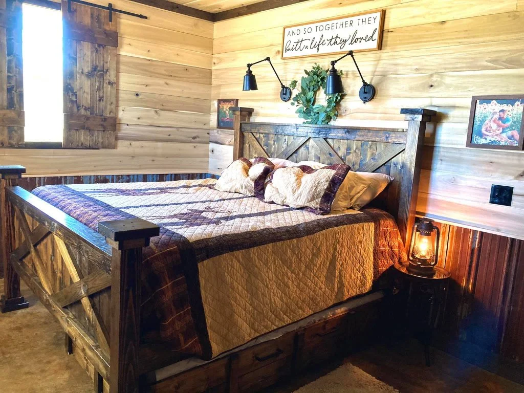 A rustic bedroom with a wooden bed as the centerpiece. The bed has a large headboard and is adorned with fluffy pillows and a cozy comforter. A wooden window is next to the bed providing natural light and a lampshade on the bedside table provides additional lighting