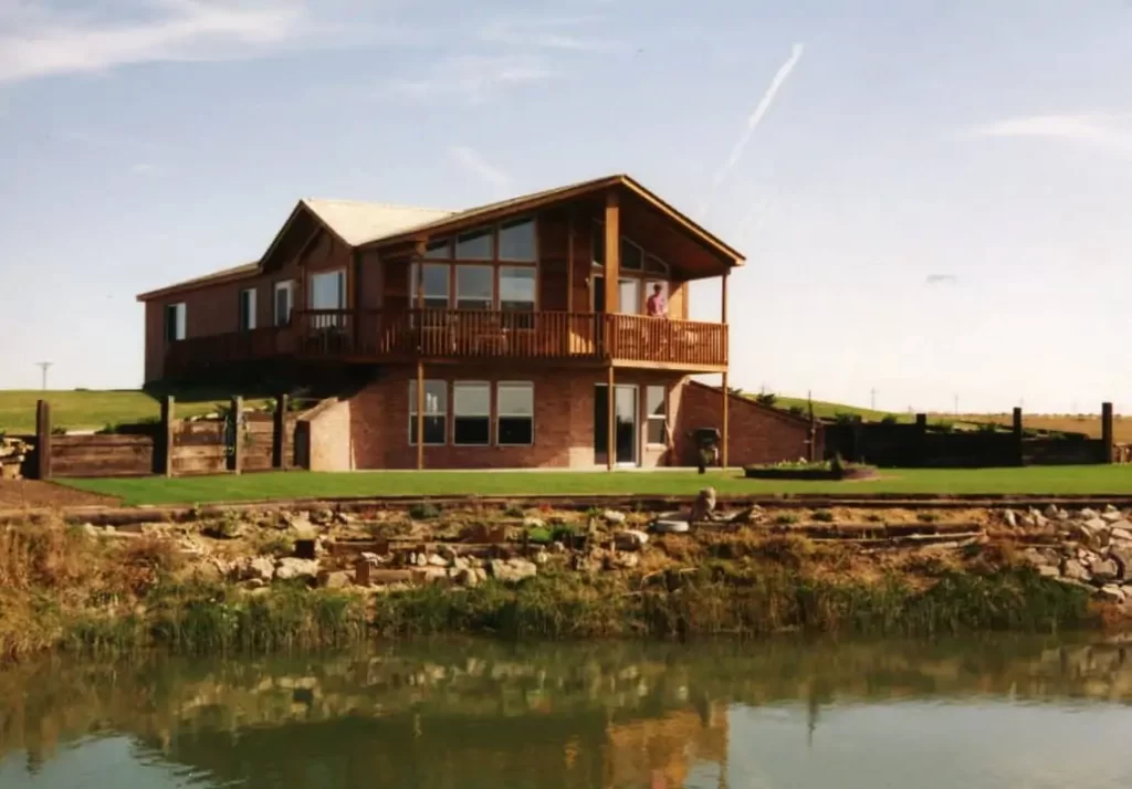 A two-story brown wooden home nestled alongside a wide lake. The front of the house features small glass windows, allowing natural light to flood the interior and providing a beautiful view of the lake. 
