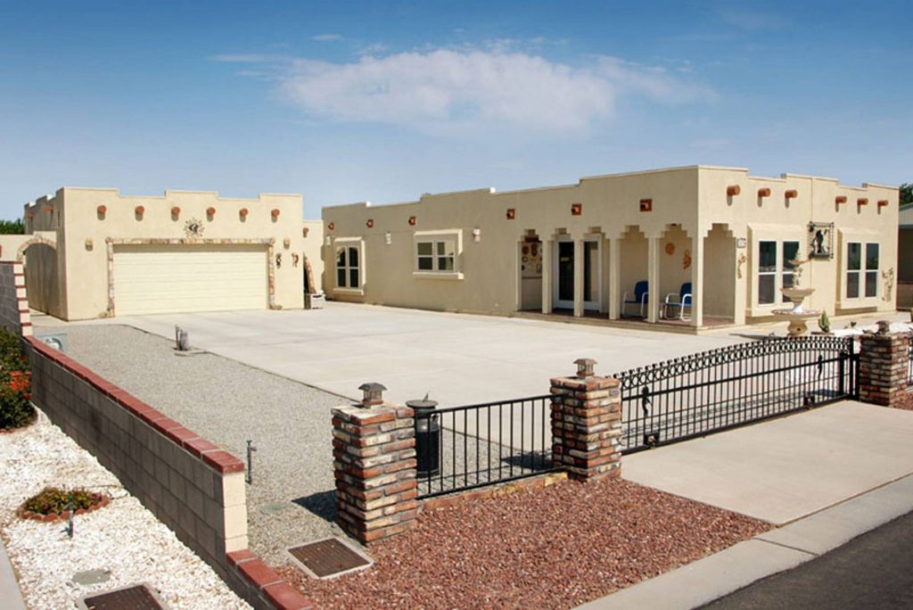 A single-story spacious home in a desert-style architecture, featuring a white exterior and a roll-down garage door. The home is surrounded by a brick fence and set against a barren desert landscape, giving it a rustic and rugged feel.