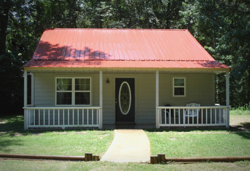 A single-story white barndominium with a gabled metal roof and a wide front porch supported by wooden columns. The building features a combination of white metal and wood siding, with large windows on either side of the entrance. In the foreground, a manicured lawn with neatly trimmed bushes and trees can be seen, while in the background, a clear blue sky provides a beautiful contrast to the white tones of the house. The overall style of the structure is modern and minimalist, with clean lines and a neutral color palette that creates a sleek and elegant look.