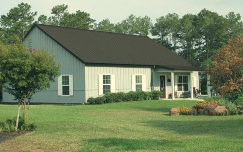 A grey barndominium with a sloping metal roof and a wide front porch with wooden columns. The building features a combination of grey metal and wood siding, with large windows on either side of the entrance. In the foreground, a gravel driveway leads up to the house, while in the background, trees and greenery can be seen framing the building. The overall style of the structure is contemporary and stylish, with a sleek and streamlined look that is complemented by the natural surroundings.