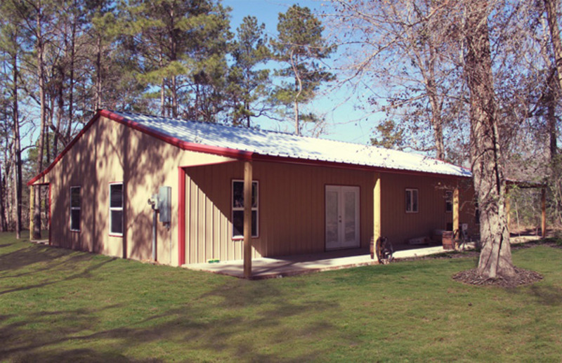 A brown barndominium with a sloping metal roof and a large front porch with wooden columns. The building features a combination of wood and metal siding, with a row of windows on the upper level and a set of double doors on the lower level. In the foreground, a gravel driveway leads up to the house, while in the background, trees and greenery can be seen framing the building. The overall style of the structure is rustic and charming, with a warm and inviting feel that is enhanced by the natural surroundings.