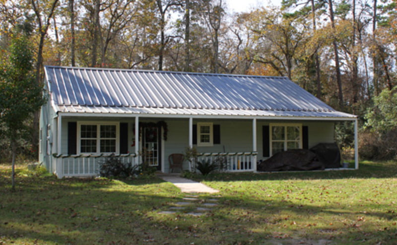 A white barndominium with a sloping metal roof and a wide front porch with wooden columns. The building features a combination of white metal and wood siding, with large windows on either side of the entrance. In the foreground, a well-manicured lawn with neatly trimmed bushes and trees can be seen, while in the background, a clear blue sky provides a beautiful contrast to the white tones of the house. The overall style of the structure is modern and chic, with a clean and minimalist look that creates a sense of elegance and sophistication.