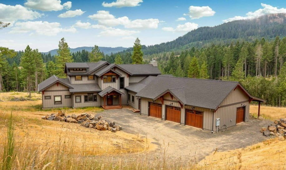 A light brown wooden barndominium is situated in a pastoral setting, surrounded by green grass and tall trees. The rustic-style house features a steeply-pitched metal roof with a chimney stack protruding from the top, and a large front porch with a white railing. The front of the home has several large windows, allowing natural light to pour in and offering picturesque views of the surrounding countryside. The overall aesthetic is cozy and inviting, perfect for a peaceful retreat away from the hustle and bustle of city life.