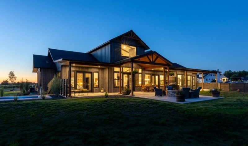 A stunning blue barndominium in a rural setting is captured in this image. The building's unique design incorporates both metal and wood materials, creating a distinctive blend of modern and traditional architecture. The structure features a large central entrance and several windows of varying sizes that allow natural light to flood the interior. The surrounding landscape is lush with greenery and mature trees, while a gravel driveway leads up to the building. The image depicts a peaceful and serene environment, perfect for those seeking a relaxing lifestyle away from the hustle and bustle of the city.