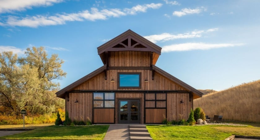 An angled view of a two-story brown barndominium is depicted in this image, nestled in the heart of a peaceful rural landscape. The exterior of the building features a mix of wood and metal materials, giving it a modern yet rustic appearance. The design is symmetrical, with a central entrance leading to the interior, and multiple windows on either side. The building's spacious driveway is surrounded by well-manicured lawns and mature trees. The image captures the serenity and beauty of countryside living while highlighting the functional and contemporary design of the barndominium.