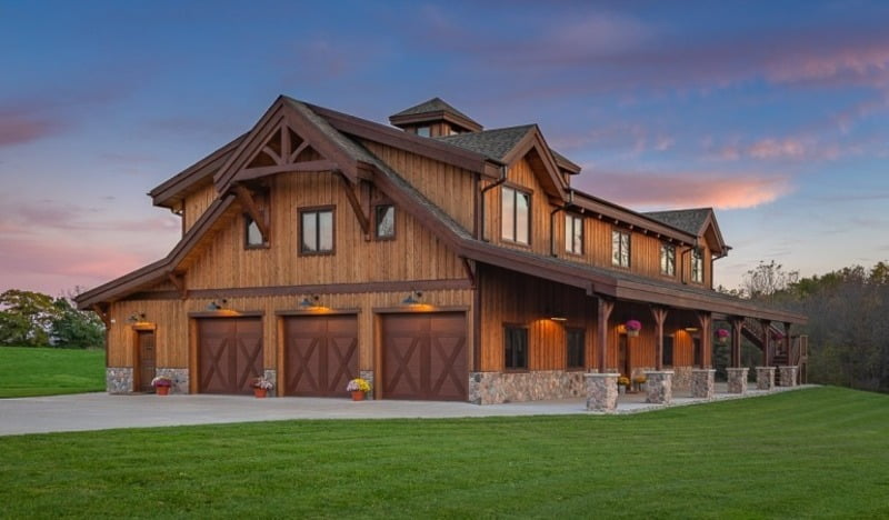An angled view of a large brown garage barndominium is depicted in this image, surrounded by a lush green countryside. The building features a modern yet rustic design, with a large central garage door and several windows for natural light to enter. The exterior is constructed with a mix of wood and metal materials, giving it a unique and contemporary appearance. The driveway leading to the building is wide and well-maintained, and the surrounding landscape is dotted with trees and open fields. The image captures the spaciousness and functionality of the garage barndominium design while showcasing the tranquility of a rural environment.