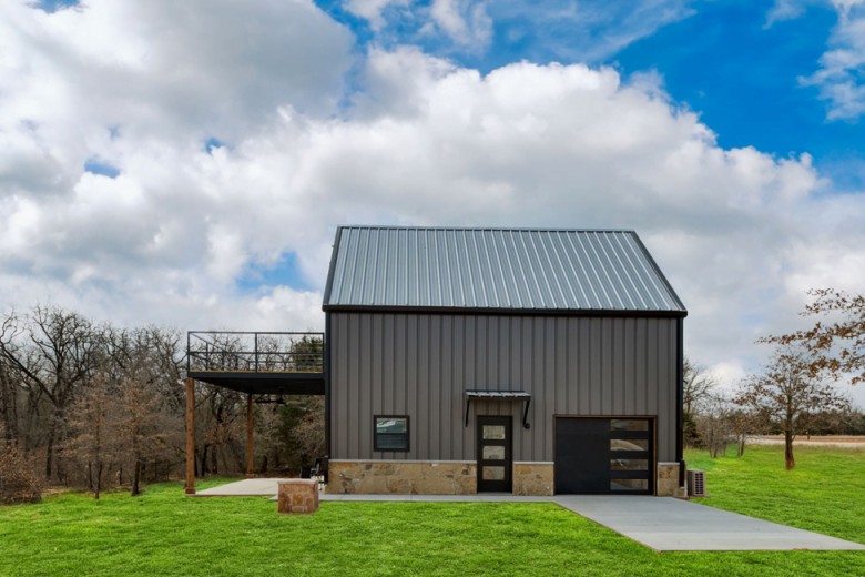 An image of a lovely single-story grey barndominium situated in a serene rural environment. The house features a grey metal exterior with white trim and roofing, giving it a clean and modern look. The front of the house has a porch with wooden railings and ample seating area, providing a perfect spot to enjoy the peaceful surroundings. The property is surrounded by green trees and a well-manicured lawn, with a gravel driveway leading up to the house. The overall aesthetic is simple yet charming, with a warm and inviting atmosphere that makes it an ideal home for anyone looking to live in the countryside.