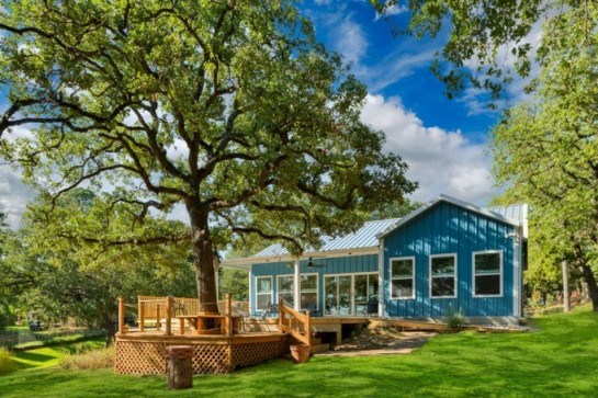 An image of a stunning blue barndominium with a lush and well-manicured lawn. The house features a blue metal exterior with white trim and roofing, giving it a refreshing and inviting look. The front of the house has a porch with wooden railings and ample seating area, providing a perfect spot to enjoy the beautiful surroundings. The property is surrounded by green trees, with a paved driveway leading up to the house. The lawn is a vibrant green and has been landscaped to perfection, with well-placed trees and bushes adding to the overall beauty of the property. The overall aesthetic is elegant and serene, with a comfortable and welcoming atmosphere that makes it an ideal home for anyone looking for a peaceful and picturesque living environment.