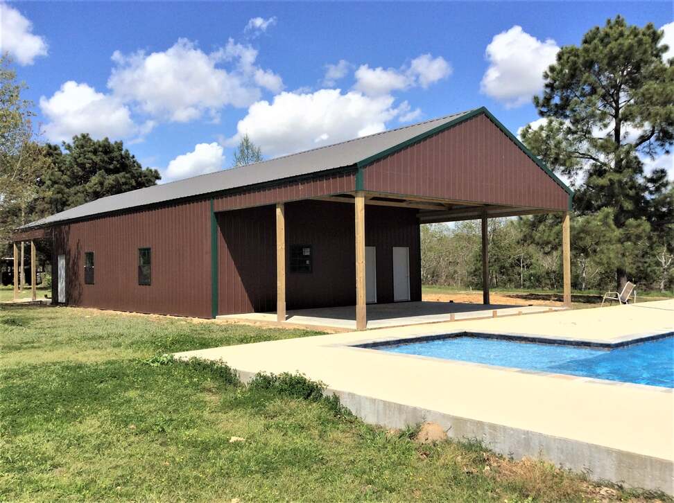 A red barndominium with a pool, featuring two white doors and surrounded by lush greenery. The building provides a stylish and relaxing rural oasis.