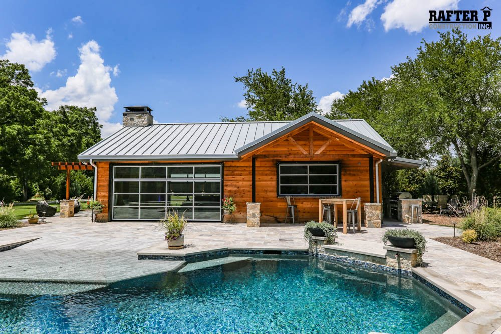A wooden cabana Barndominium is surrounded by a shimmering blue pool. The building boasts large glass windows that offer stunning views of the surrounding area and enhance its natural, rustic aesthetic.
