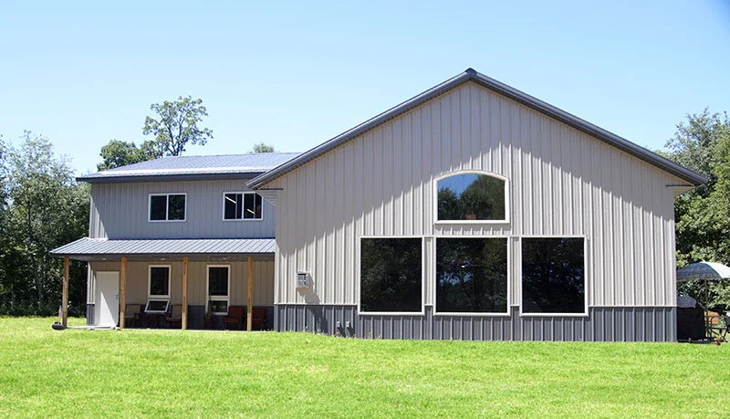 An image of a stunning white and grey two-story barndominium, featuring a metal roof, and large windows on both levels. The exterior of the building is surrounded by a lush green lawn, with beautiful trees and bushes adding to the serene atmosphere. The modern design of the building is complemented by the rustic aesthetic of the barndominium, making it a perfect blend of contemporary and traditional architecture.