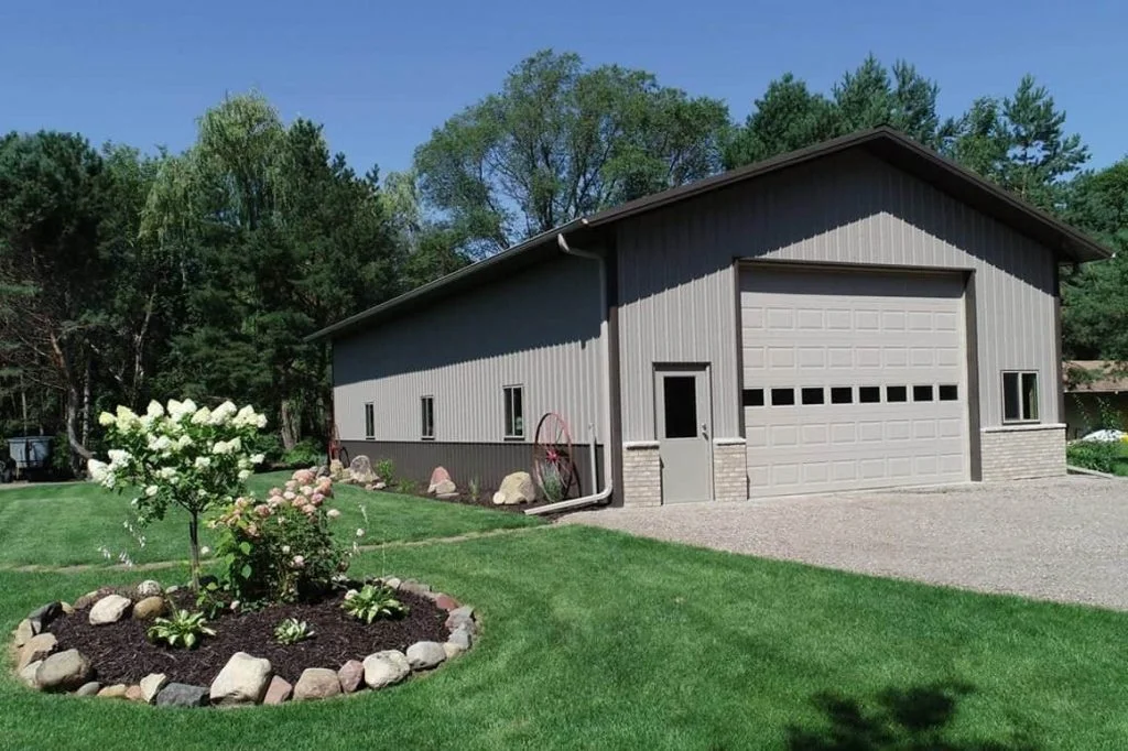 An image of a sleek and modern grey storage barndominium situated in a rural area. The building features a tall, gabled metal roof with grey metal siding and black trim. Two large garage doors flank the front of the building, providing access to the expansive storage space inside. The surrounding land is flat and open, with fields and hills visible in the distance. A gravel driveway leads up to the building, with a few trees and shrubs providing a natural touch to the landscape.
