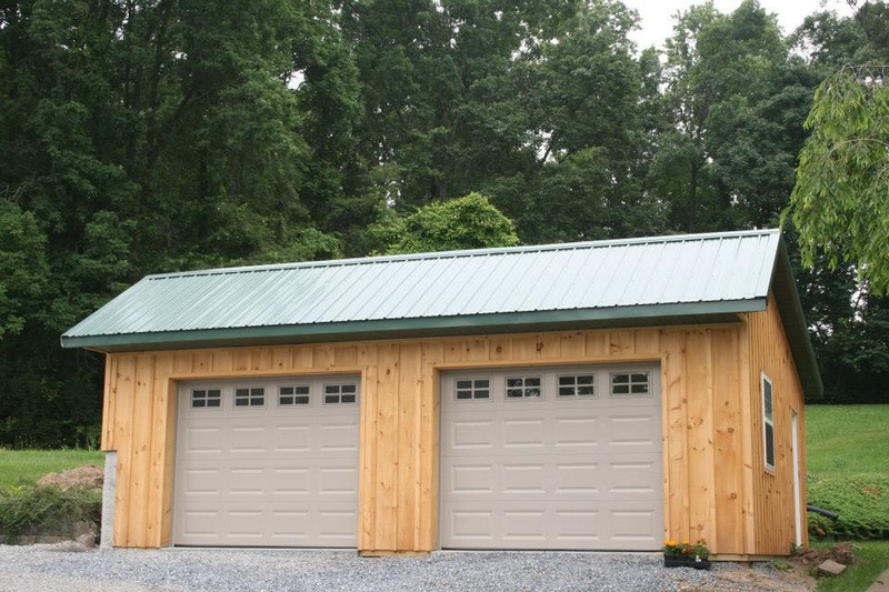An image of a charming wooden barndominium with two grey garage doors and a green roof. The building's natural wood exterior and rustic design blend well with the surrounding landscape, evoking a sense of warmth and comfort. The two grey garage doors add a functional and practical touch to the building, while the green roof provides a pop of color and an eco-friendly element. This may be a residential or commercial space, catering to the needs of a rural community or a nature lover.