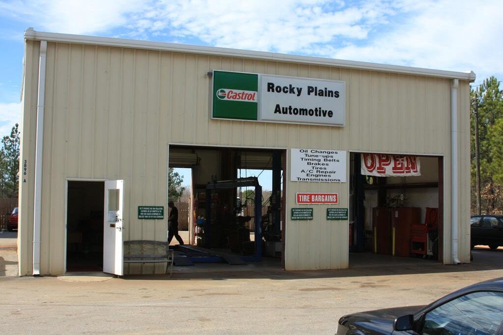 An image featuring a peach-colored commercial metal building that serves as an auto shop. The building has a rectangular shape and a flat roof, with several large windows on the front and sides. The entrance to the auto shop is a large garage door on the front of the building, with a smaller entrance door next to it. The exterior walls are made of metal panels, and there are several cars parked in front of the building. The surroundings include a paved parking lot, a few trees, and a clear blue sky in the background.