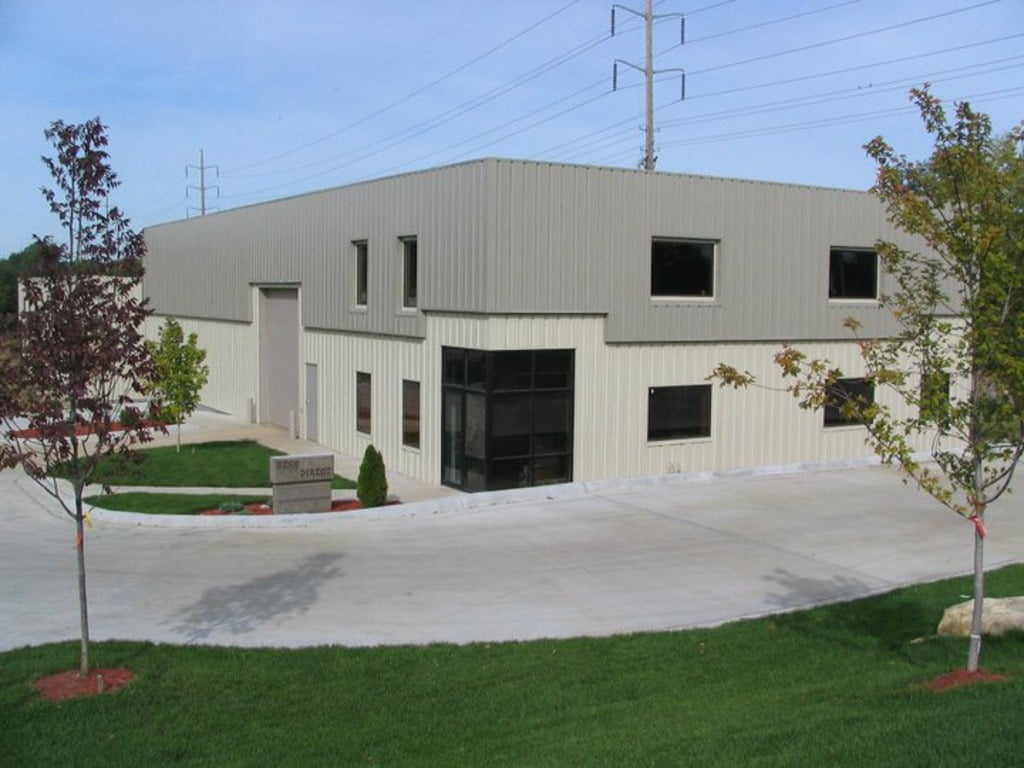 An image featuring a large grey and white industrial metal building. The building has a rectangular shape and a flat roof, with several large windows and a double-door entrance on the front. The exterior walls are made of metal panels, and there are no visible signs of any landscaping or other structures in the immediate surroundings. The image is set against a clear blue sky background, and the building's size and appearance suggest that it may be a manufacturing plant, warehouse, or other similar industrial facility.