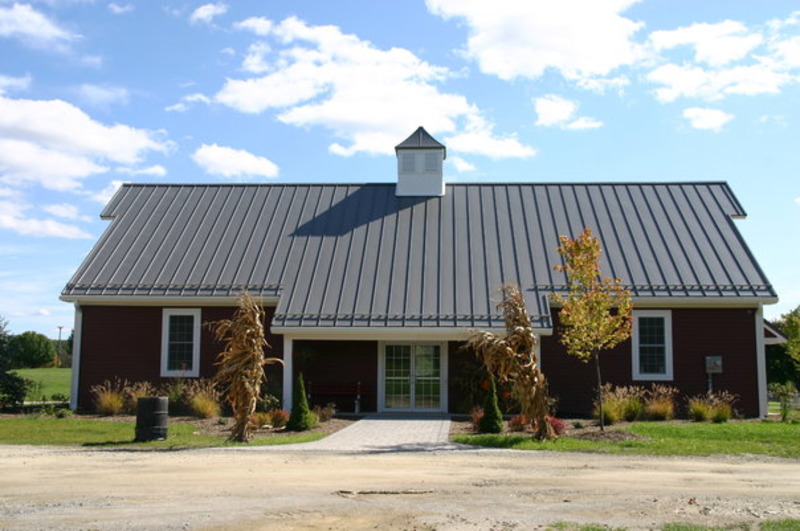 An image of a brown barndominium with a sleek grey roof. The building features two small windows and a central glass door, allowing ample natural light inside. The solid brown color of the building creates a warm and inviting atmosphere