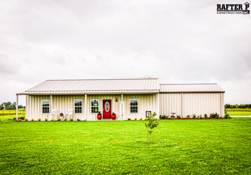 A simple white Barndominium sits on a wide, lush green lawn. The building features a small glass window on the side and a vibrant red door, adding a pop of color to its clean and modern design.