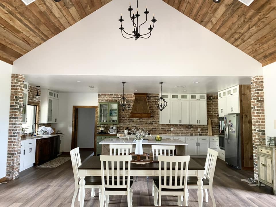 A cozy and rustic kitchen with a white color scheme. The photo is taken from the perspective of a dining table in the foreground, which has a wooden top and white chairs