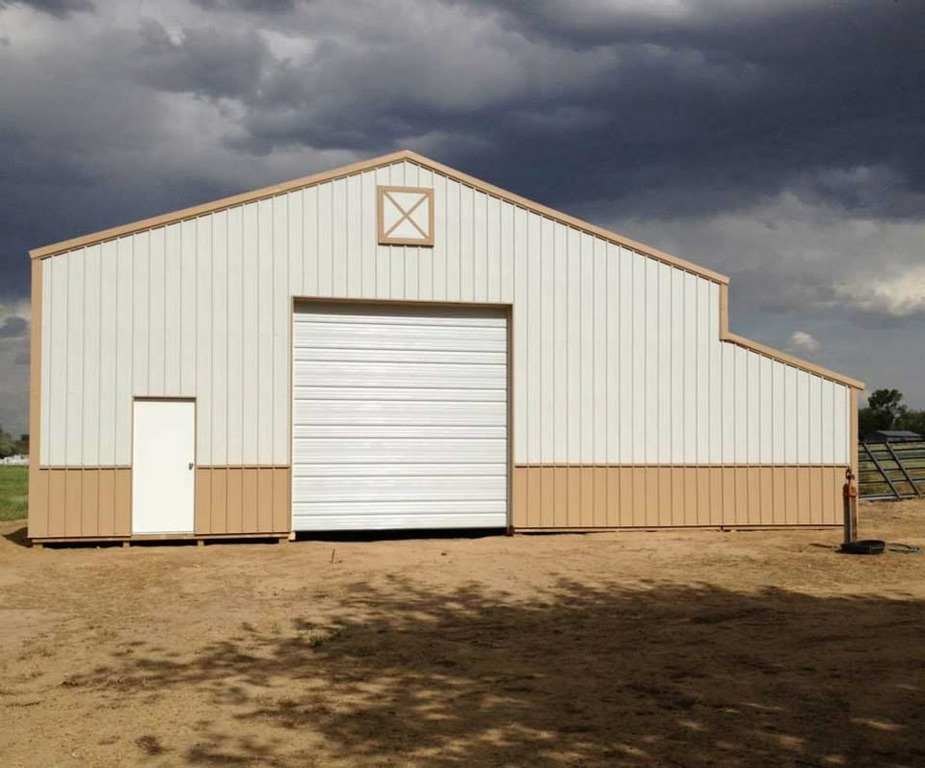 An image of a spacious metal horse barn in white and brown colors, set against a lush green landscape. The barn has a sloped metal roof with a brown finish, and the exterior walls are in white metal siding with large sliding barn doors. The barn features several horse stalls with wooden doors and windows, a tack room for storing horse equipment, and an open area for grooming and training. The surrounding area is dotted with mature trees, a paddock with a white fence, and a beautiful blue sky above, making it a perfect haven for horses and their owners.