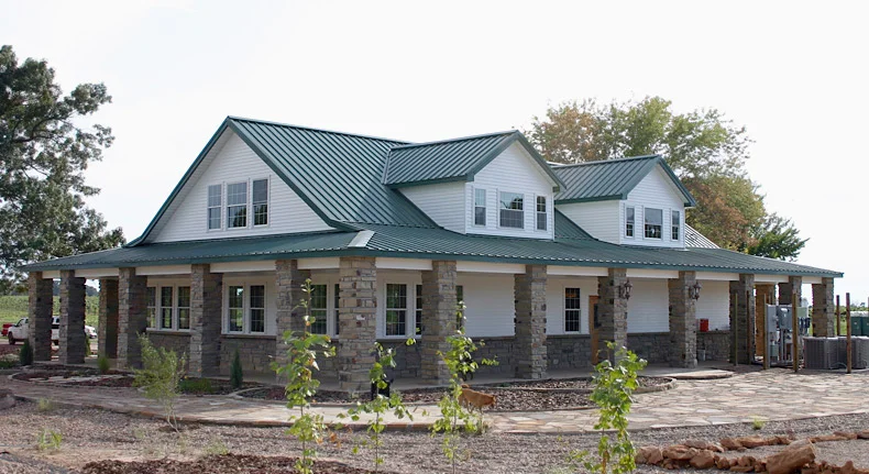 An image of a modern white Kodiak steel home located in Arkansas, set against a bright blue sky. The house is a single-story rectangular structure with a gable roof, and the exterior walls are made of light-colored steel panels with a smooth surface. The front of the house features large floor-to-ceiling windows that let in plenty of natural light, and a small covered porch with a single entry door. The surrounding area is mostly flat, with sparse vegetation and no other buildings in sight, suggesting that the house is located in a rural area.