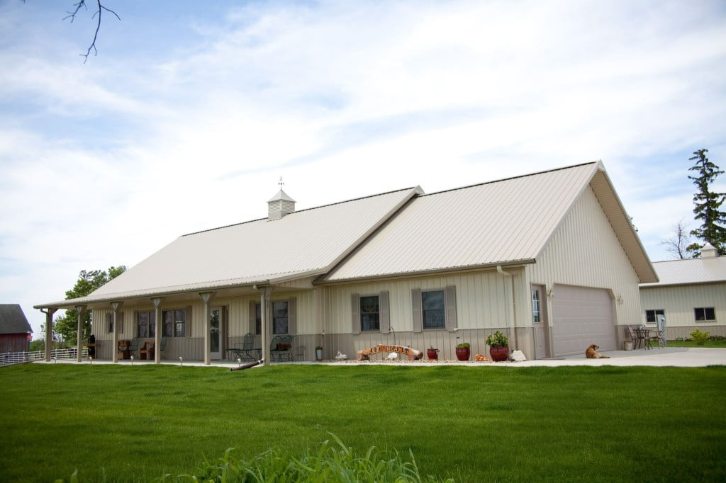 A spacious light brown barndominium featuring a large central entrance with a gable roof and sliding glass doors. The building has a symmetrical design with a row of windows on either side of the entrance and a metal roof that complements the natural wood siding. The surrounding area is open with a few scattered trees and green grass in the foreground. The image captures the peaceful and rustic ambiance of a modern barndominium with its clean lines, warm color, and natural surroundings.