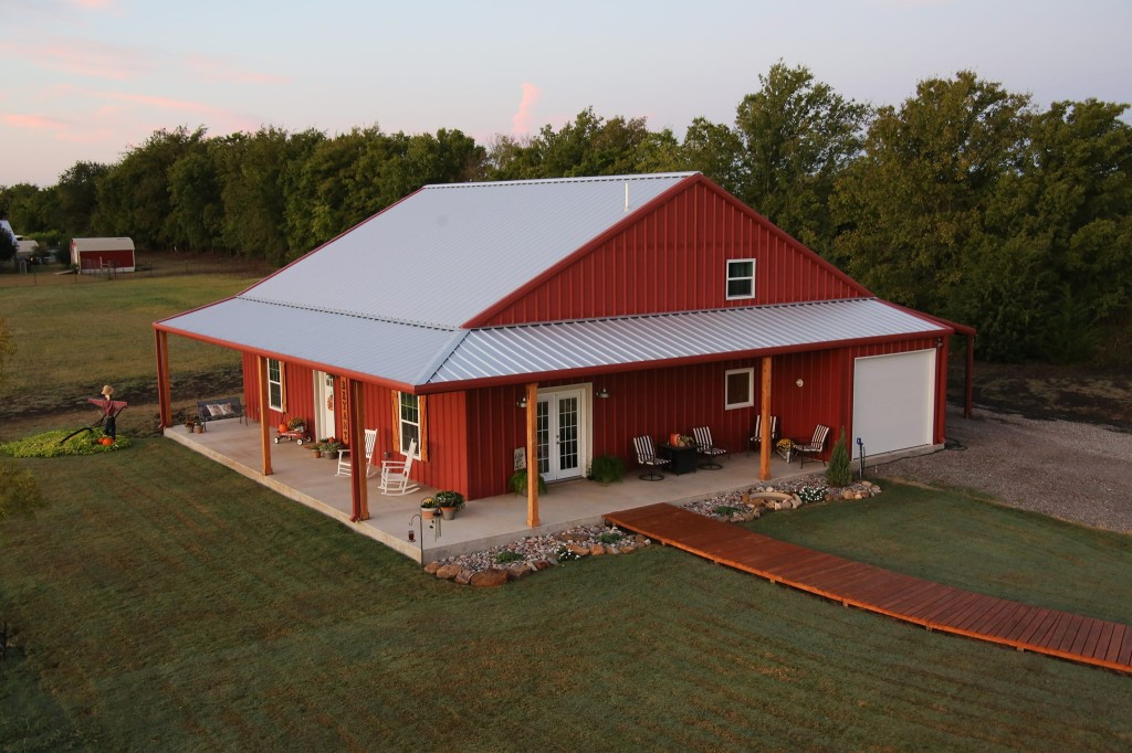 An image of a red Barndominium built by Mueller Inc, located in Texas. The building is a rectangular structure with a gabled roof, and the exterior is made of durable steel panels painted in a vibrant red color. The bottom portion of the building features a rustic red brick facade, adding to its unique and charming appearance. The entrance to the building is located on one of the shorter sides, and there are several windows of varying sizes on both sides of the structure. The surrounding area is mostly flat, with some sparse vegetation and a few other buildings visible in the background, suggesting that the structure is located in a rural or suburban area.