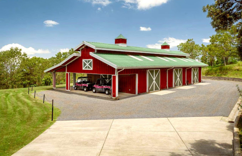 An image of a striking red Barndominium, standing out against a beautiful natural landscape. The building has a sloped metal roof with a bold red finish and a spacious front porch with wooden railings and a swing. The exterior walls are a combination of red metal panels and light-colored wood siding, with large windows providing plenty of natural light. The entrance features a sliding barn door with a natural wood texture, adding a rustic touch to the modern design. The surrounding area has a mix of lush greenery, a pond, and rolling hills, enhancing the beauty and charm of the red Barndominium."