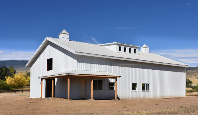 An image of a charming grey Barndominium, featuring a combination of modern and rustic design elements. The building has a sloped metal roof with a deep grey finish and a wrap-around porch with wooden posts and railings. The exterior walls are a mix of grey metal panels and light-colored wood siding, and the entrance has a sliding barn door with a modern steel frame. Large windows provide ample natural light and offer stunning views of the surrounding greenery. The surrounding landscape has a well-manicured lawn, mature trees, and shrubs, complementing the Barndominium's warm and inviting aesthetic.