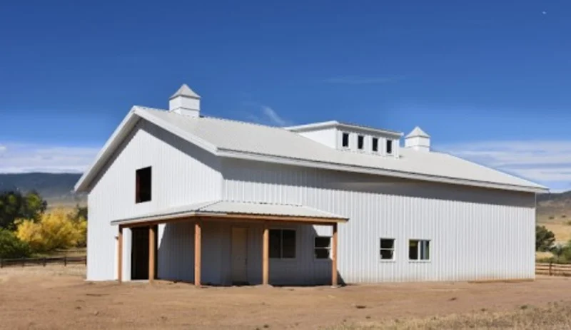 An image of a white Barndominium built by Sunward Steel Buildings, located in Denver. The building is a unique rectangular structure with a sloped roof, and the exterior is made of durable steel panels painted in a bright white color. The bottom portion of the building features a rustic red brick facade, adding to its charming and unique character. The entrance to the building is located on one of the shorter sides, and there are several windows of varying sizes on both sides of the structure. The surrounding area is mostly flat, with some sparse vegetation and a few other buildings visible in the background, suggesting that the structure is located in a rural or suburban area