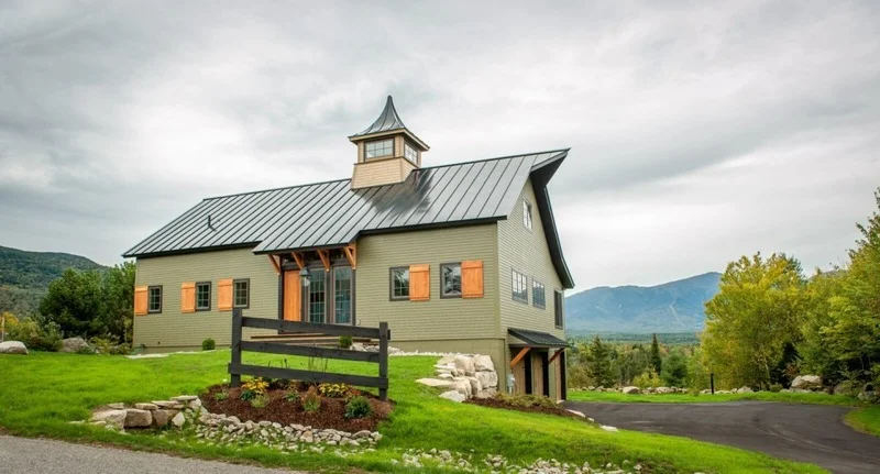 An image of a stylish grey barndominium, a unique residential building that combines the rustic design of a barn with the modern comfort of a home. The building features a symmetrical design with a sloping metal roof, grey wooden siding, and large windows that allow plenty of natural light into the living spaces. The surrounding landscape includes green grass, trees, and a gravel driveway leading up to the house. The grey color of the building's exterior creates a sleek and modern look, while the warm sunlight gives it a cozy and inviting feel. This beautiful and serene scene exudes a sense of simplicity and elegance.