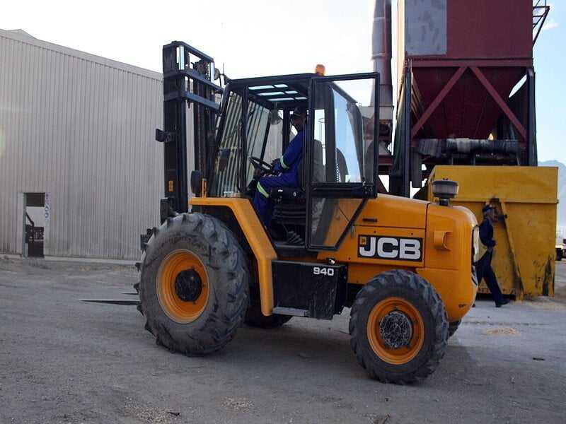 A sturdy yellow rough terrain forklift with chunky tires and a robust frame, navigating a challenging terrain strewn with rocks and uneven ground. 