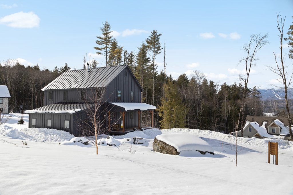 A prefab home that's been beautifully covered in snow and is being complimented by tall trees that surrounds it. Image provided by huntingtonhomesvt.com