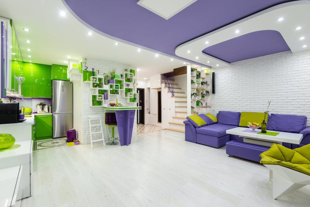 A boldly-designed green, white and purple-themed kitchen with a white floor, white and green walls, and a white and purple ceiling. A purple and yellow couch is next to a staircase at the entrance of the kitchen.