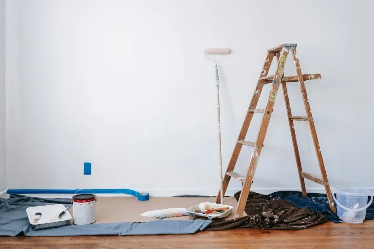 Home improvement is in progress showing a freshly painted white wall. used paintbrushes, a paint can, a ladder, and other painting paraphernalia are on the ground.
