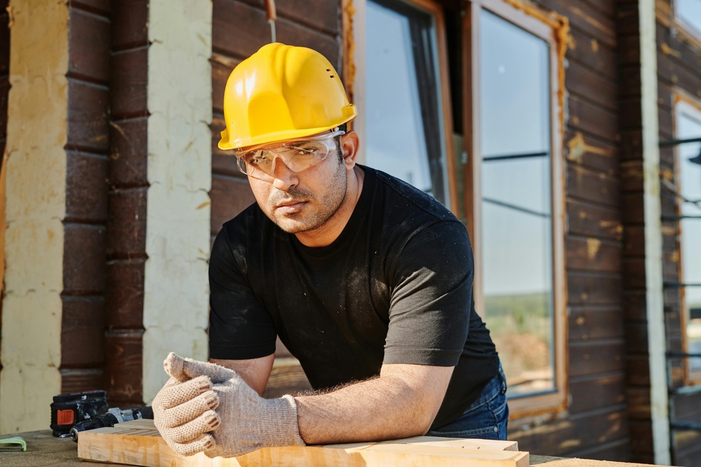 A contractor in a black T-shirt and blue jeans wearing safety glasses and a yellow helmet 