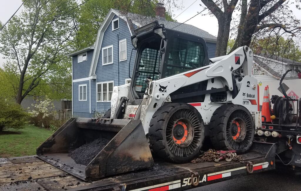 The Bobcat S770 skid steer loader loaded on the back of a truck with a blue house and trees in the background